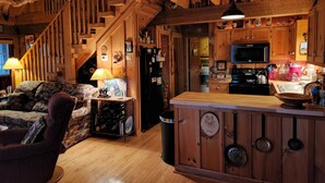 A cozy cabin interior with a wooden staircase, a living area with a sofa and table, a kitchen with a stove and microwave, and a small island with hanging pans. - Beautiful Sunsets & Amazing Stars in every season, Family-owned gem (Meadows of Dan)