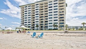 A multi-story hotel with balconies, a beach area with chairs and umbrellas, and palm trees.