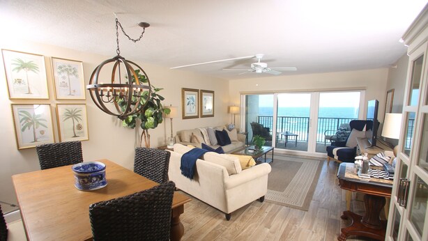 A spacious living area with a large dining table, comfortable seating, a ceiling fan, and a view of the ocean through large sliding doors.