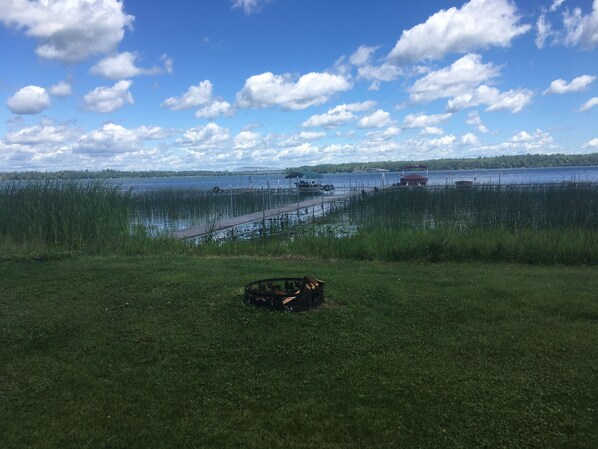A grassy area with a dock extending into a lake, surrounded by reeds and trees in the background.