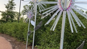 Three metallic wind sculptures with reflective surfaces and illuminated elements, set in a landscaped area with greenery and a dirt path. - The Blue Door Bentonville is an ideal Bike In/Bike Out location, close to dwtwn. (Bentonville)