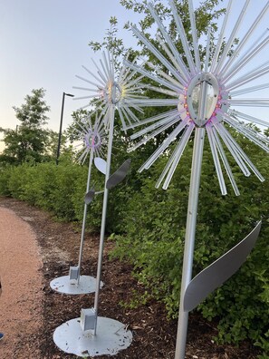 Three metallic wind sculptures with reflective surfaces and illuminated elements, set in a landscaped area with greenery and a dirt path.