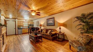 A spacious living area with wooden floors and a ceiling fan, a cozy seating area with a sofa and armchairs, and a kitchen in the background.