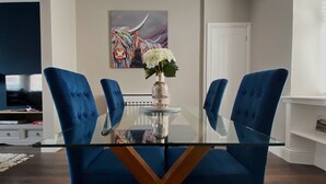 Un coin repas avec une table en verre, des chaises recouvertes d’un tissu bleu, un vase rempli de fleurs et un tableau coloré accroché au mur.