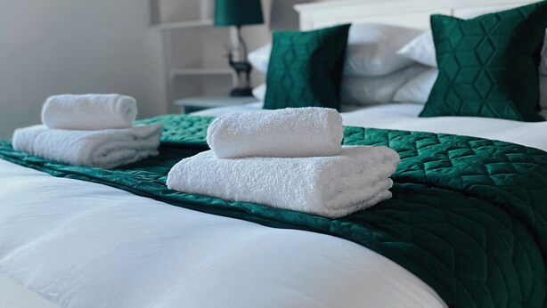 Un lit bien fait, avec des draps blancs et une couverture verte, une pile de serviettes pliées et un coussin vert posé sur une tête de lit blanche.