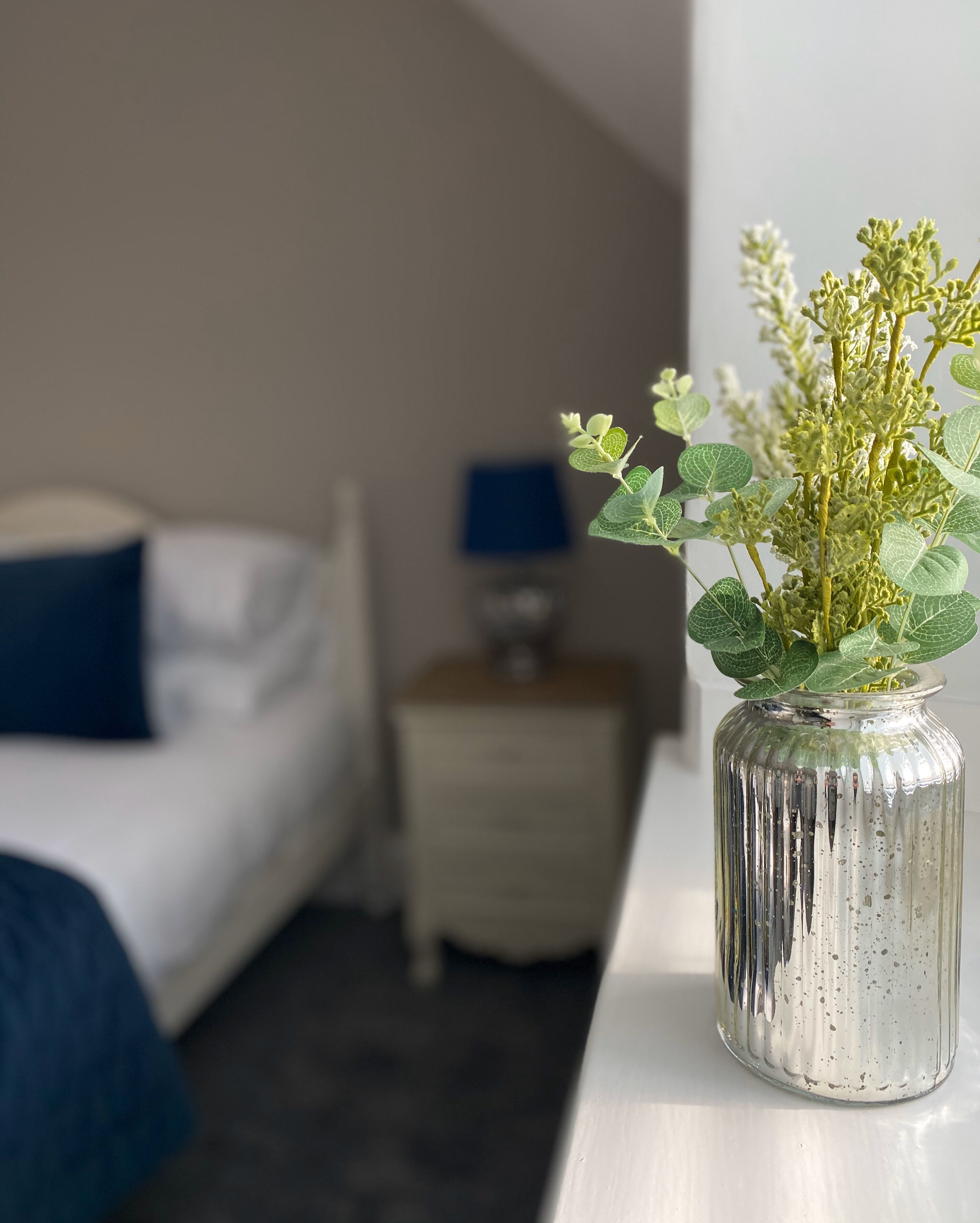 Une chambre d’hôtel avec un lit, une table de chevet, une lampe et un vase contenant des plantes vertes.