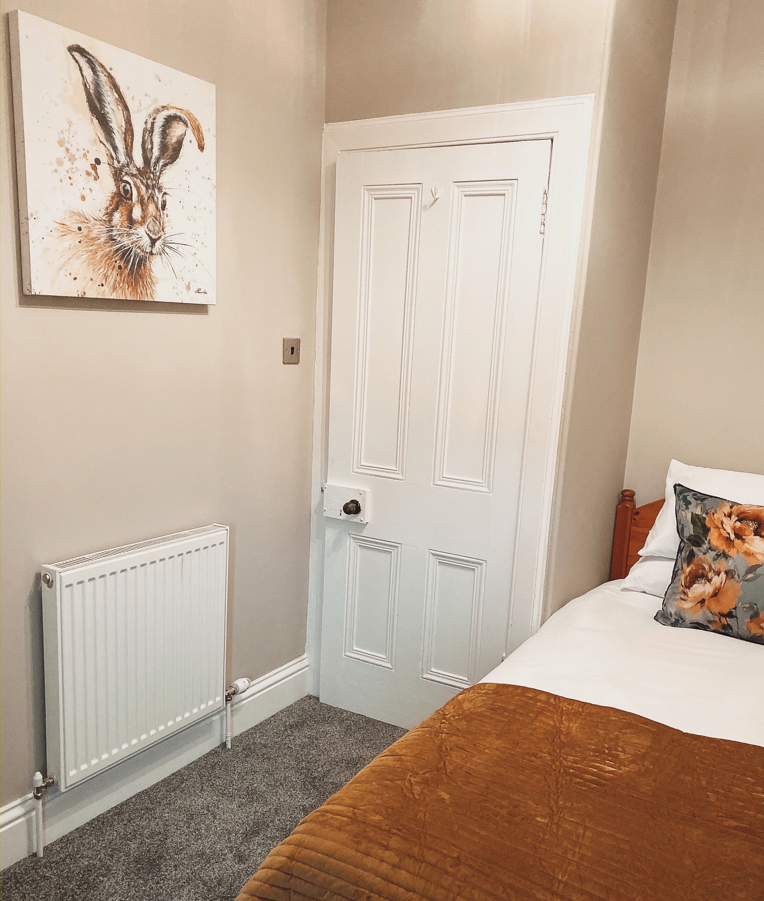 Une chambre avec une porte blanche, un radiateur, un lit recouvert d’une couverture marron et un coussin à motifs floraux.