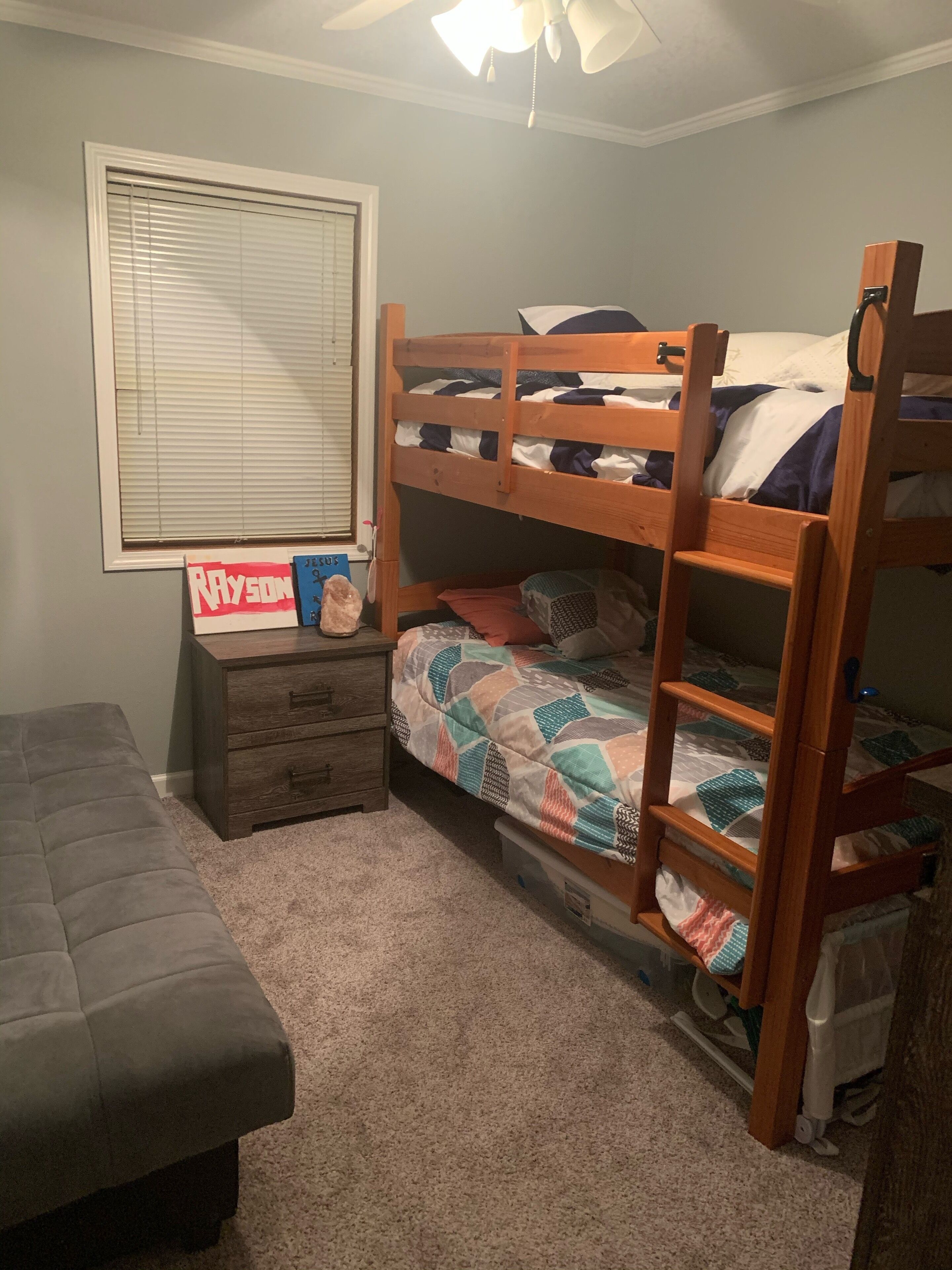 A bunk bed room with a desk and drawers, a window with blinds, and a ceiling fan.