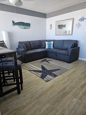 Un salon avec un canapé d’angle gris, un tapis à motifs d’étoile de mer et des œuvres d’art murales comprenant des décorations de poissons et de chevaux de mer.