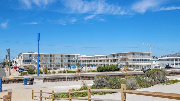 Un hotel costero con varios balcones, un estacionamiento y una playa de arena al frente.