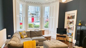 A living room with a sofa, ottoman, and a television. There is a bookshelf and a fireplace. - Meadows Sanctuary-Perfect base for exploring Edinburgh, private  garden (Edinburgh)