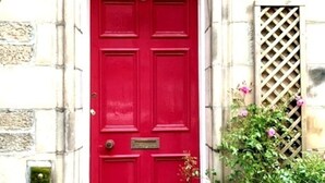 A red door with a white frame, a small window with a lattice design, and a green plant with pink flowers to the right. - Meadows Sanctuary-Perfect base for exploring Edinburgh, private  garden (Edinburgh)