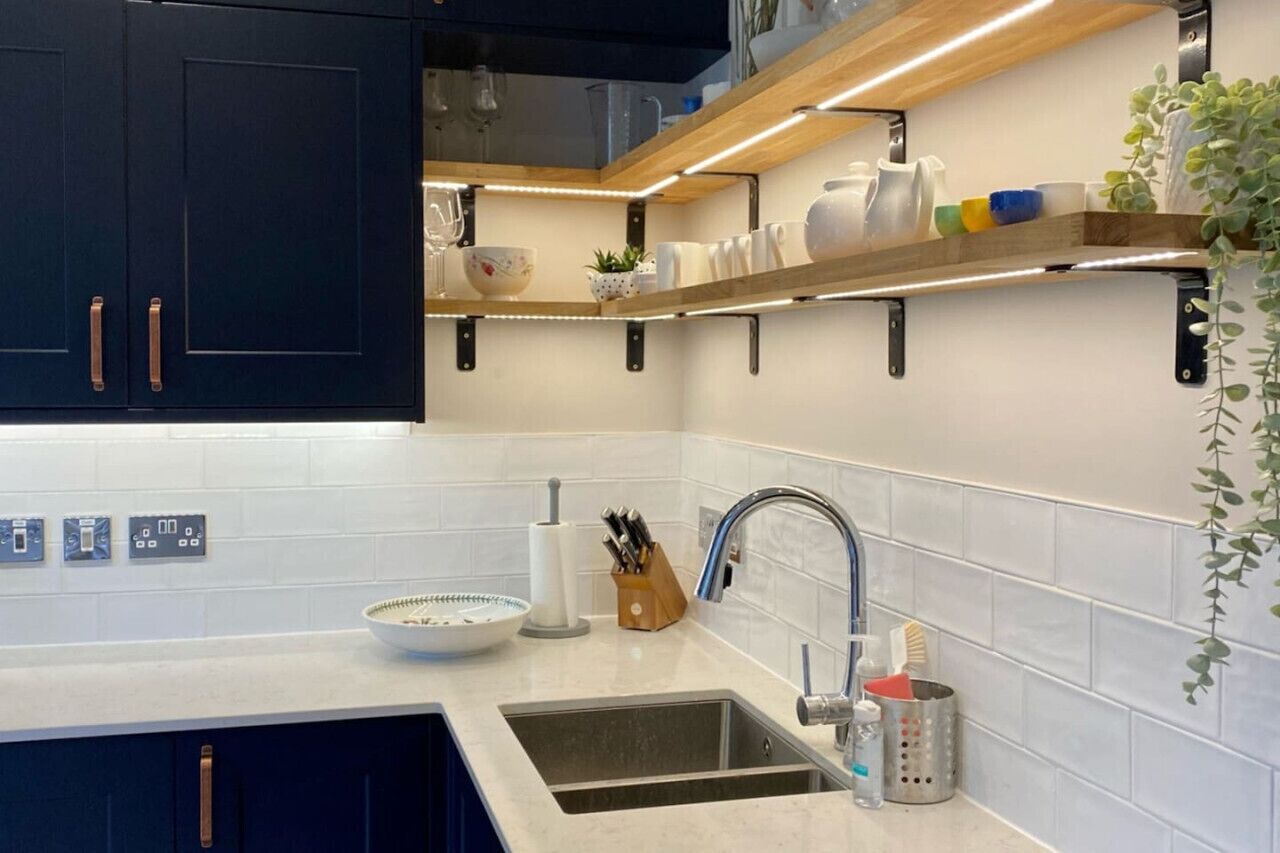 A modern kitchen with dark cabinets, white countertops, and a built-in sink. There are shelves above the counter holding various kitchen items and a plant on the right.