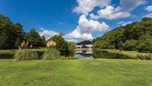 A serene park with a pond, trees, and a building in the background. - Charming  Cottage at Willow Creek Falls & Vineyard in Blue Ridge, GA (Blue Ridge)