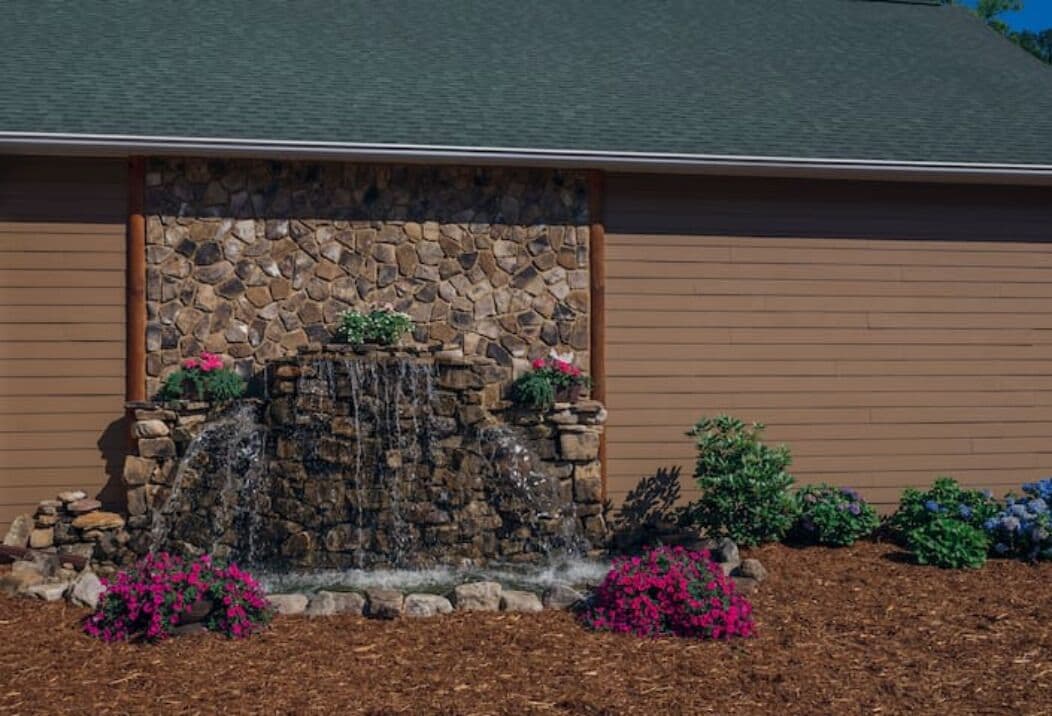 A building with a stone waterfall feature, surrounded by colorful flowers and shrubs.
