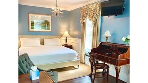 Una habitación de hotel con cama, escritorio, sillón verde, candelabro y un cuadro en la pared.