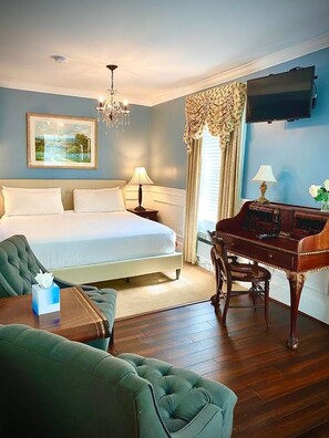 Una habitación de hotel con cama, escritorio, sillón verde, candelabro y un cuadro en la pared.