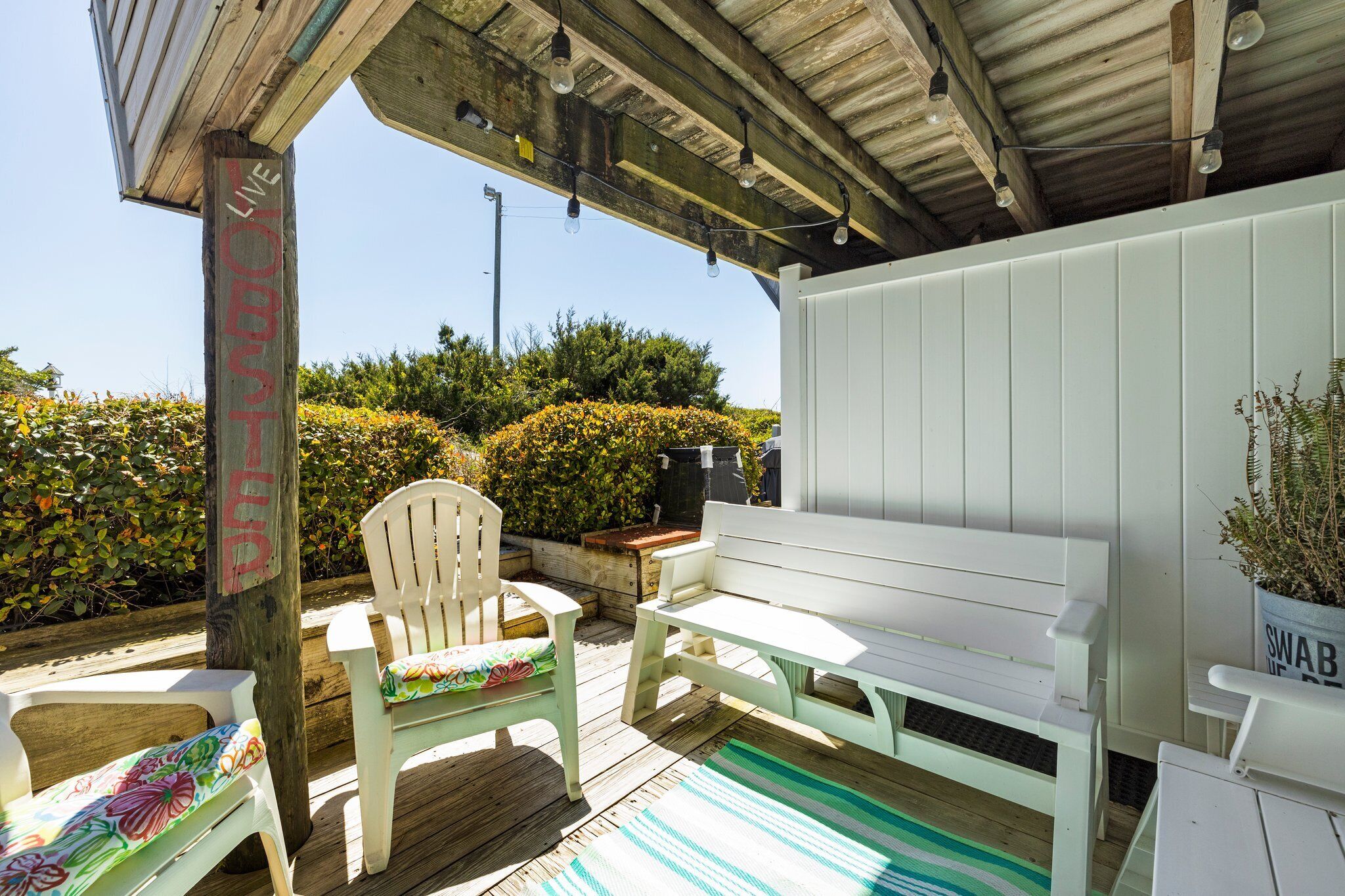 A covered outdoor seating area with a wooden deck, white furniture, and a sign that reads "LIVE ON STAND."