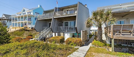 En fastighet som ligger vid stranden, med en träveranda, en trappa som leder upp till en balkong och utsikt över havet.