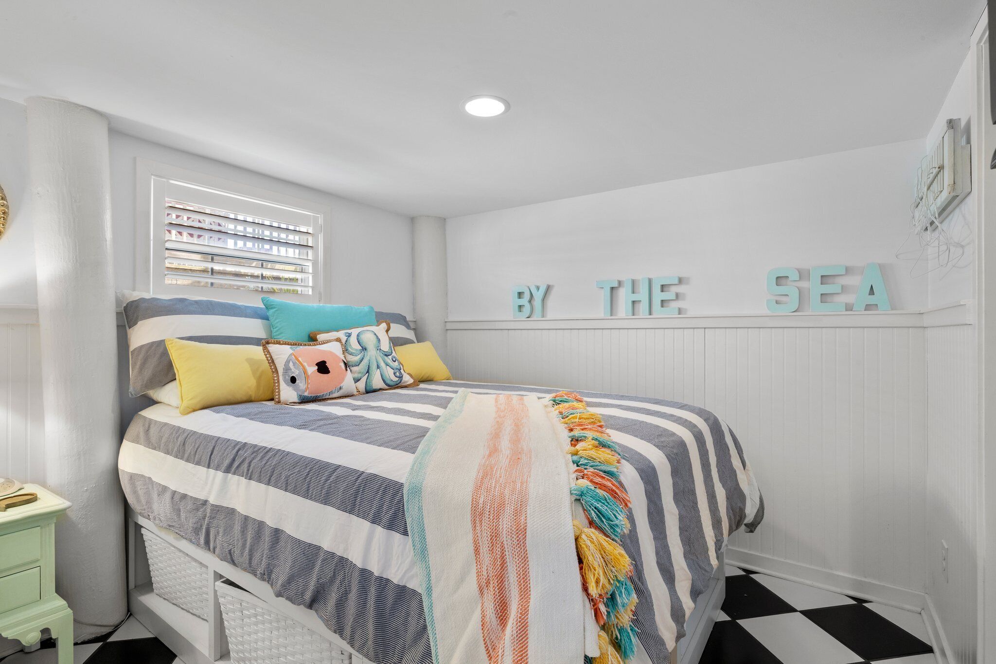 A bedroom with a bed, a nightstand, a window with blinds, and a wall sign that reads "BY THE SEA".