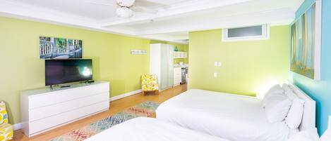 A bedroom with a bed, a TV, a ceiling fan, and a kitchen in the background.