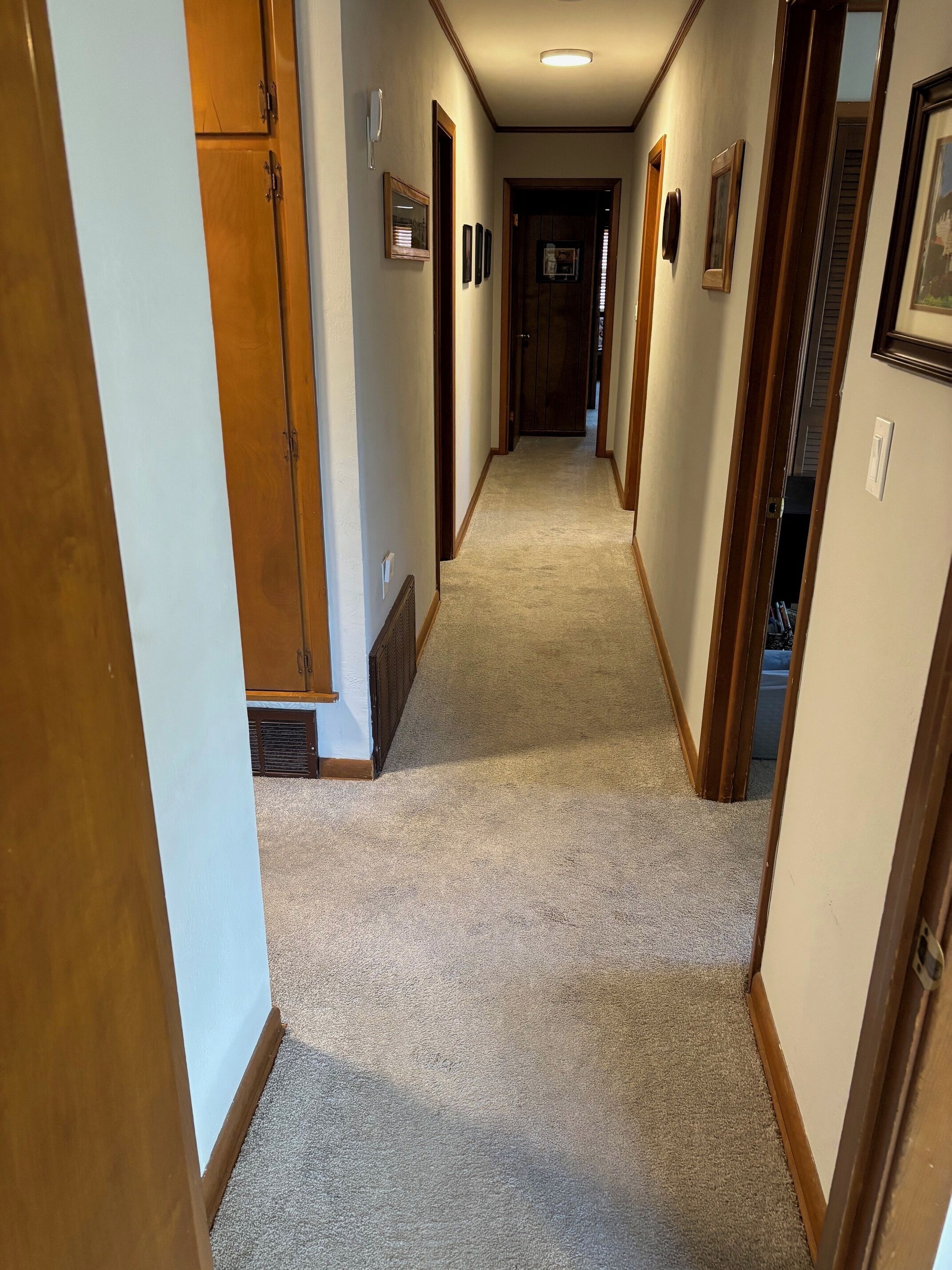A narrow hallway with carpeted floors, wooden doors, and framed pictures on the walls.