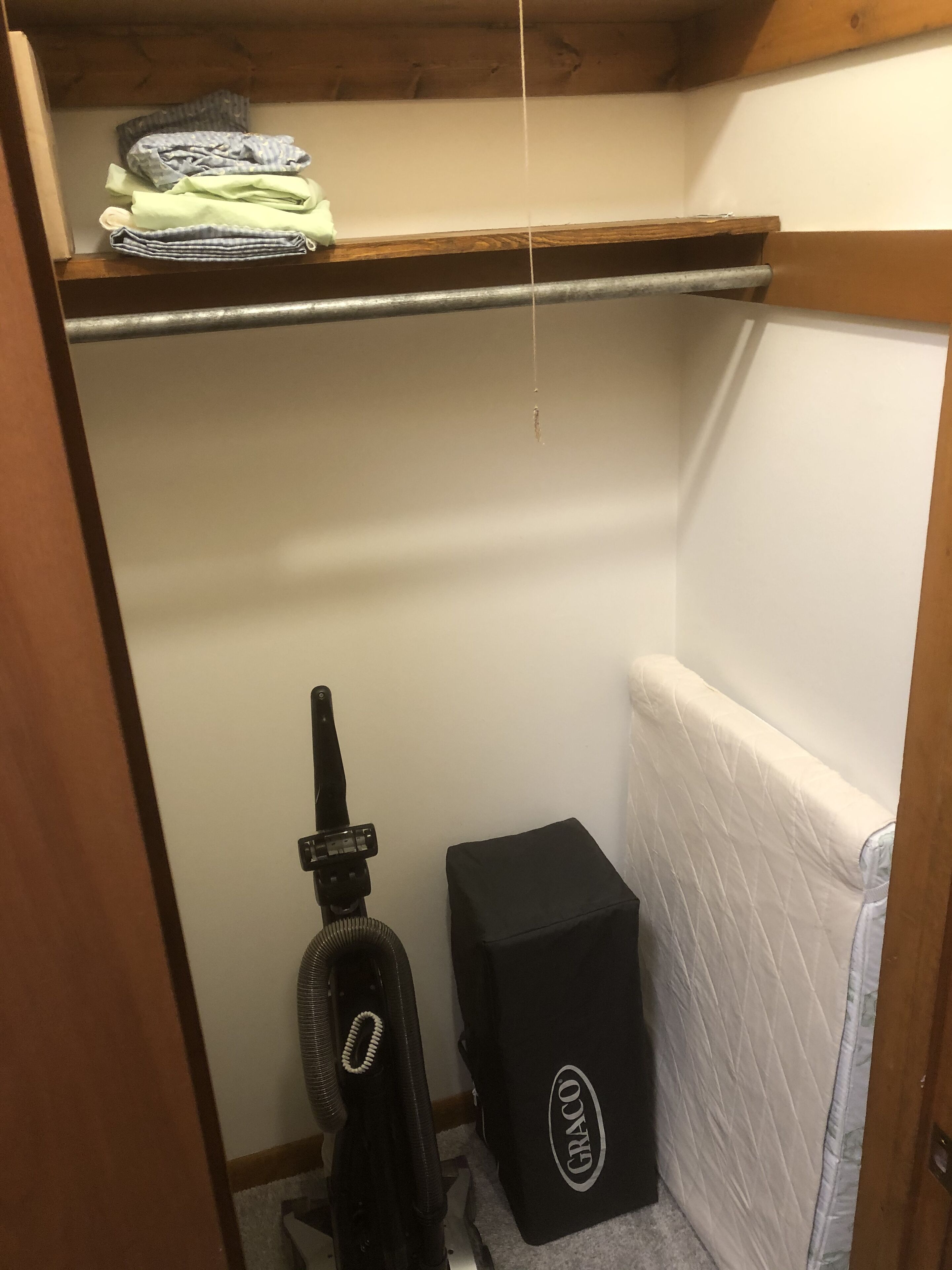 A small, narrow room with a wooden ceiling, a radiator, a mattress, a vacuum cleaner, and a folded blanket on a shelf.
