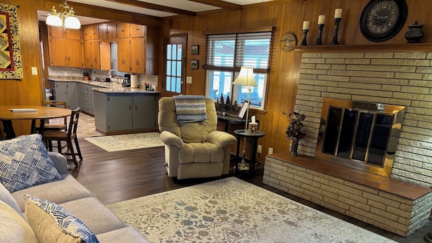 Een gezellige woonkamer met een bank, fauteuil en open haard. In de achtergrond is een keuken met houten keukenkastjes en een eethoek met tafel en stoelen te zien.