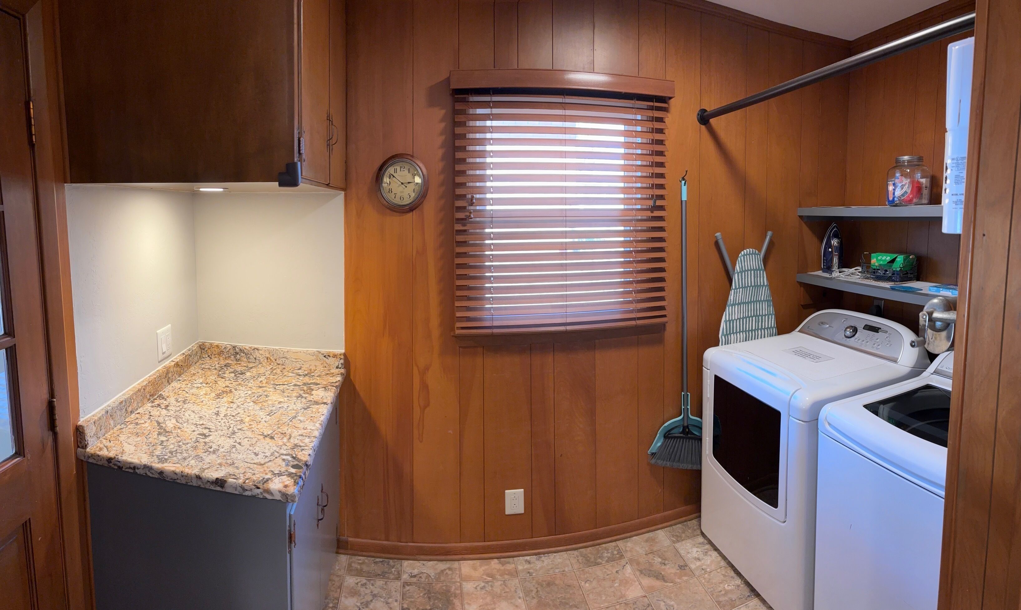 A compact laundry room with a built-in washer and dryer, wooden cabinetry, a clock, and a window with blinds.