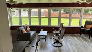 Een zonnedroom met houten vloer, een bank, een tafel en twee stoelen. Grote ramen bieden uitzicht op een achtertuin met een hek en gazon.