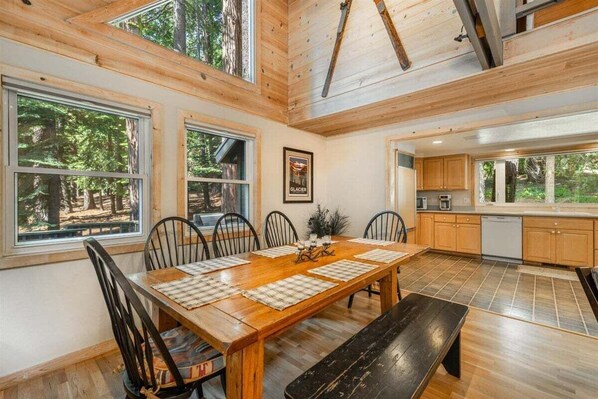 Comedor de madera con una mesa grande y sillas, zona de cocina y ventanas amplias que dan al bosque.