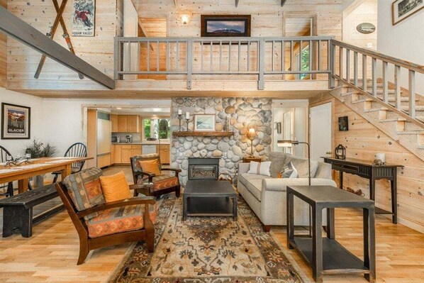 A spacious living area with a stone fireplace, wooden furniture, a dining table, and a staircase leading to an upper level.