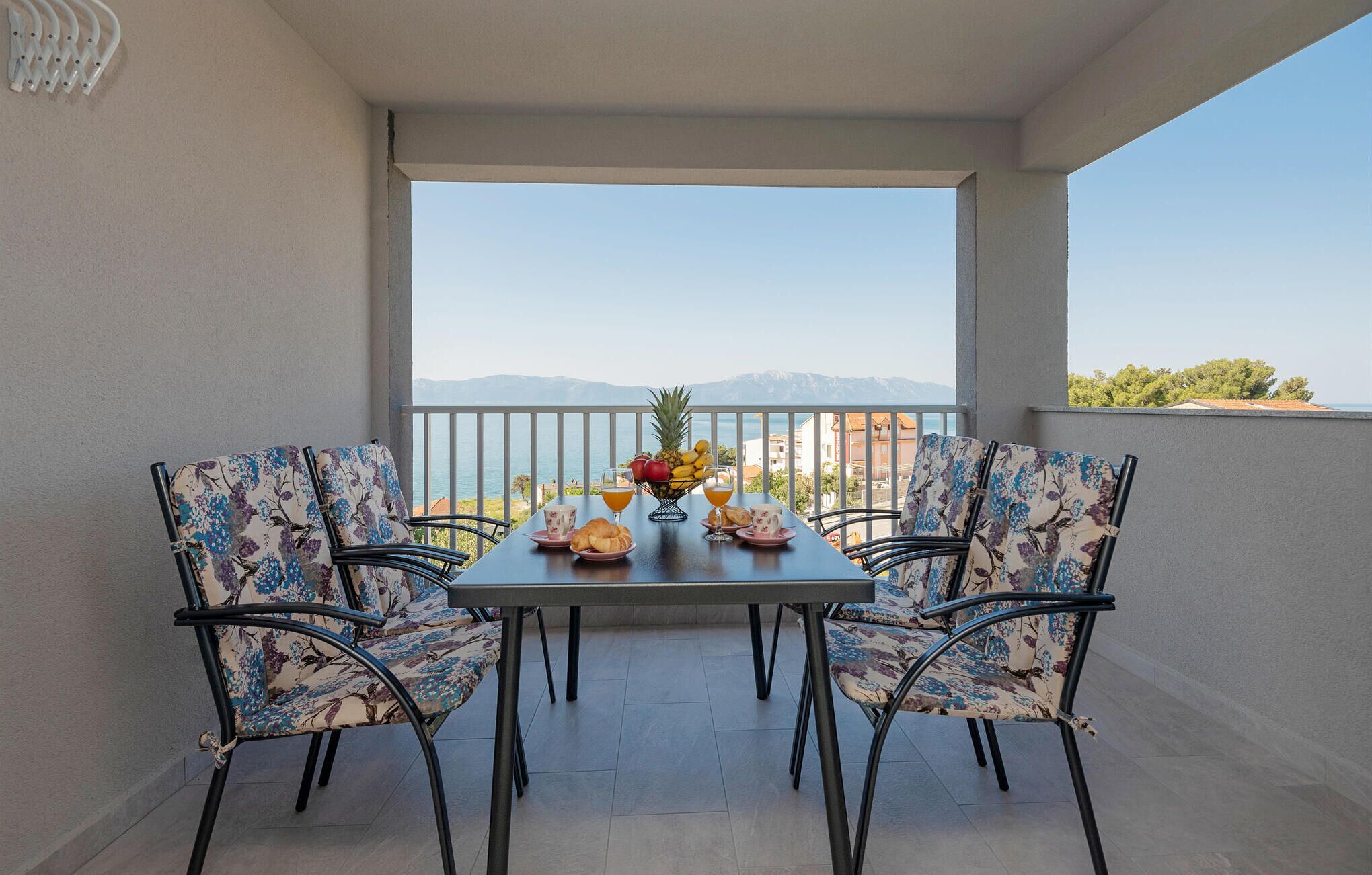 A balcony with a table set for two, chairs with floral upholstery, a fruit bowl, and a view of the sea and mountains.
