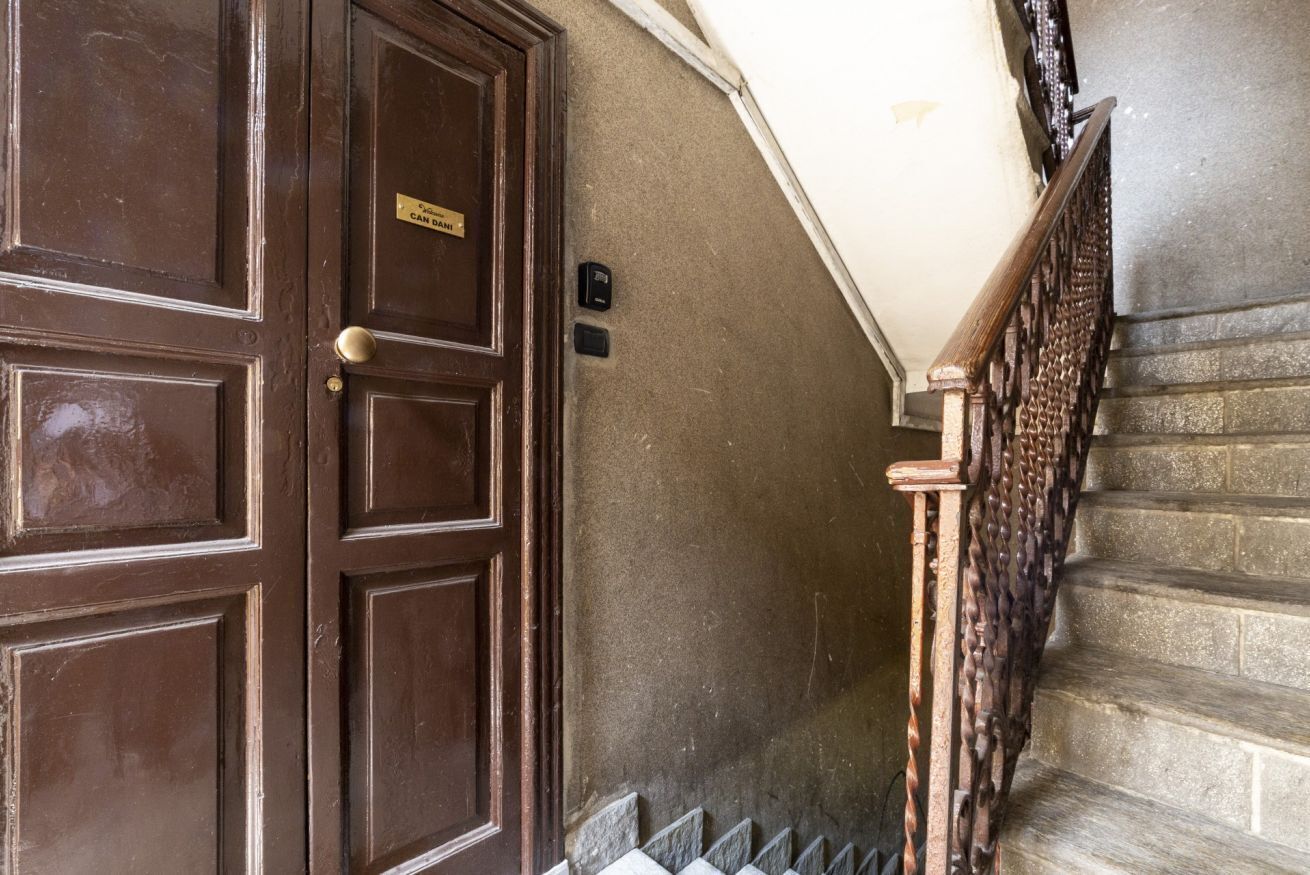 Una porta in legno con maniglia in ottone e una targa, una scala in pietra con ringhiera in legno e un muro dalla superficie irregolare.