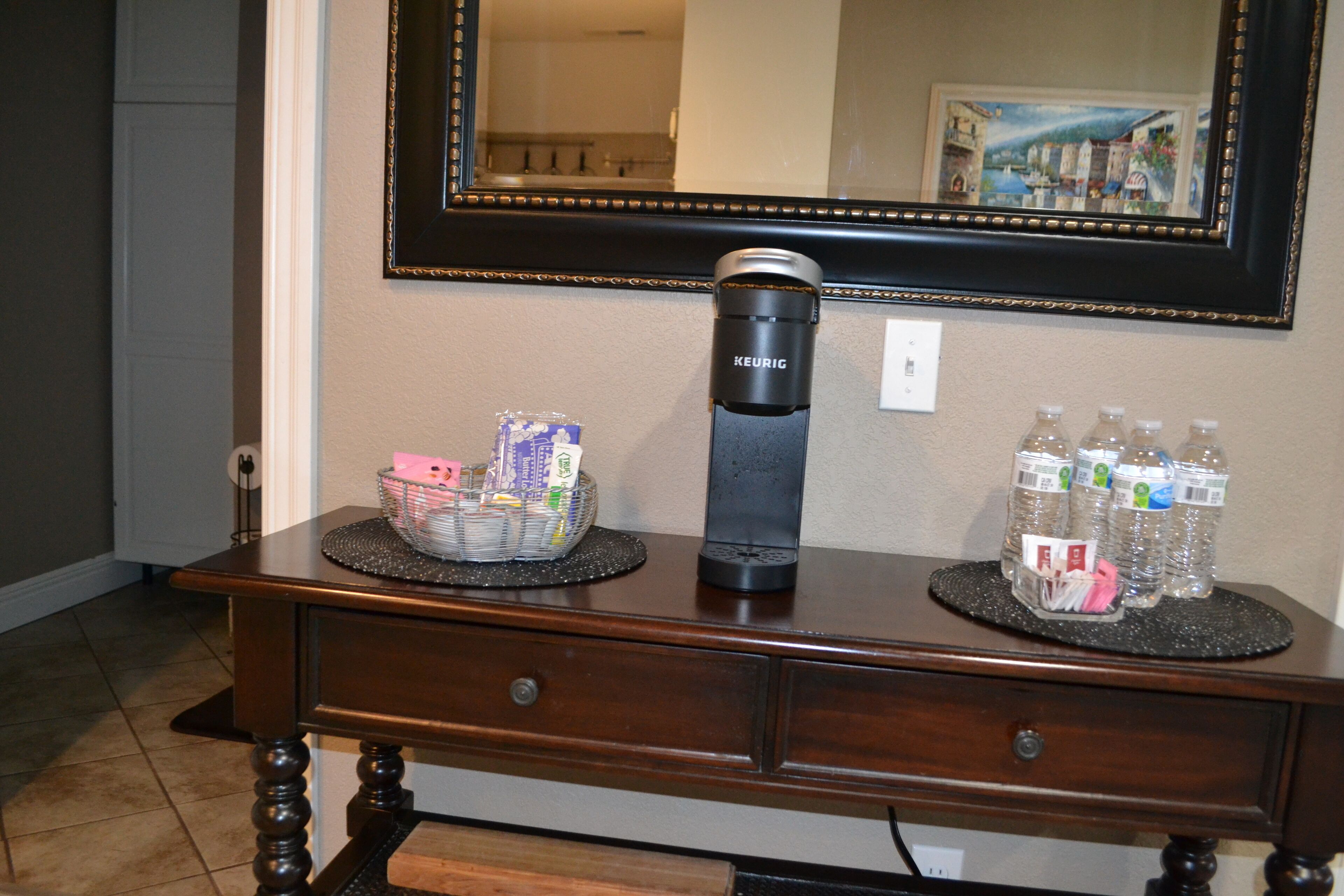 A wooden console table with a coffee maker, a basket of snacks, and bottles of water.