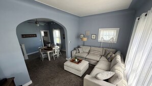 A living room with a large L-shaped sofa, a dining table with chairs, and a kitchen area in the background.