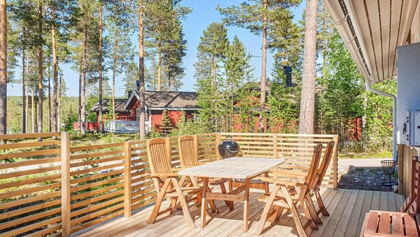 A wooden deck with a table and chairs, surrounded by a wooden fence, overlooking a forest and a red building. - 3 bedroom stunning home in Särna (Särna)