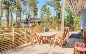 A wooden deck with a table and chairs, surrounded by a wooden fence, overlooking a forest and a red building. - 3 bedroom stunning home in Särna (Särna)