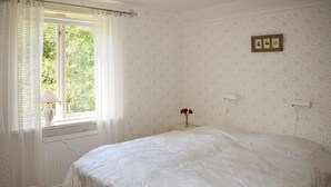 Un dormitorio con cama, una ventana con cortinas, una mesita de noche con un florero y un cuadro en la pared.