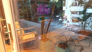 Une vue depuis une fenêtre, avec une chaise et une plante à l’intérieur. Dehors, on aperçoit une rue avec des voitures et un bâtiment doté d’une terrasse.