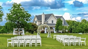 A wedding ceremony setup with white chairs arranged in rows on a grassy lawn, a floral arch, and a large house in the background. - Where the Missouri sky wakes up first.  Peaceful mornings you’ll never forget. (Warrensburg)