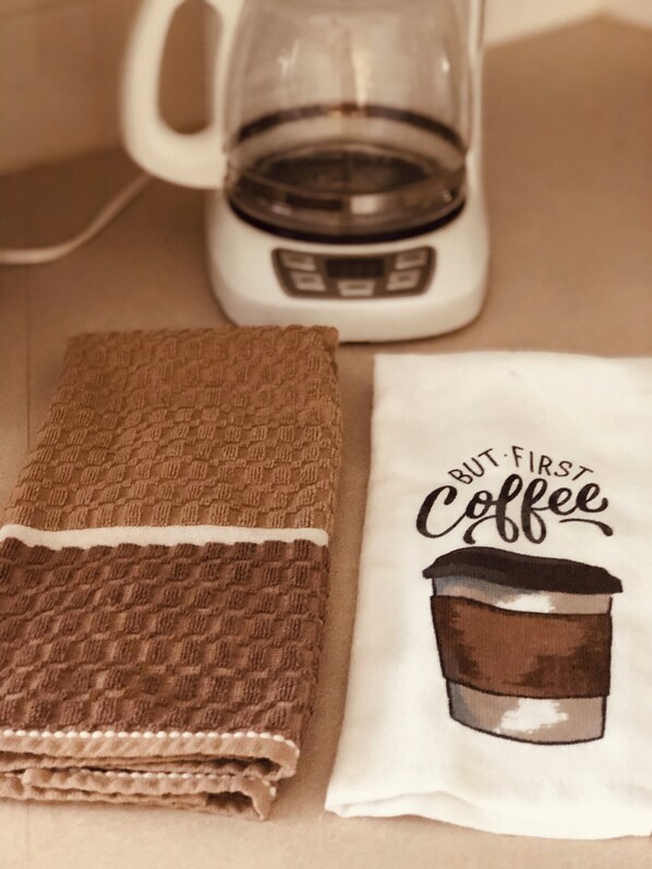 Una cocina con cafetera, una toalla marrón doblada y un paño blanco con una ilustración de taza de café y texto.