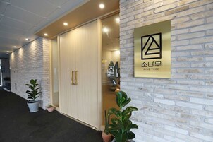 A modern building entrance with a white brick facade, a sliding glass door, and a logo on the wall.