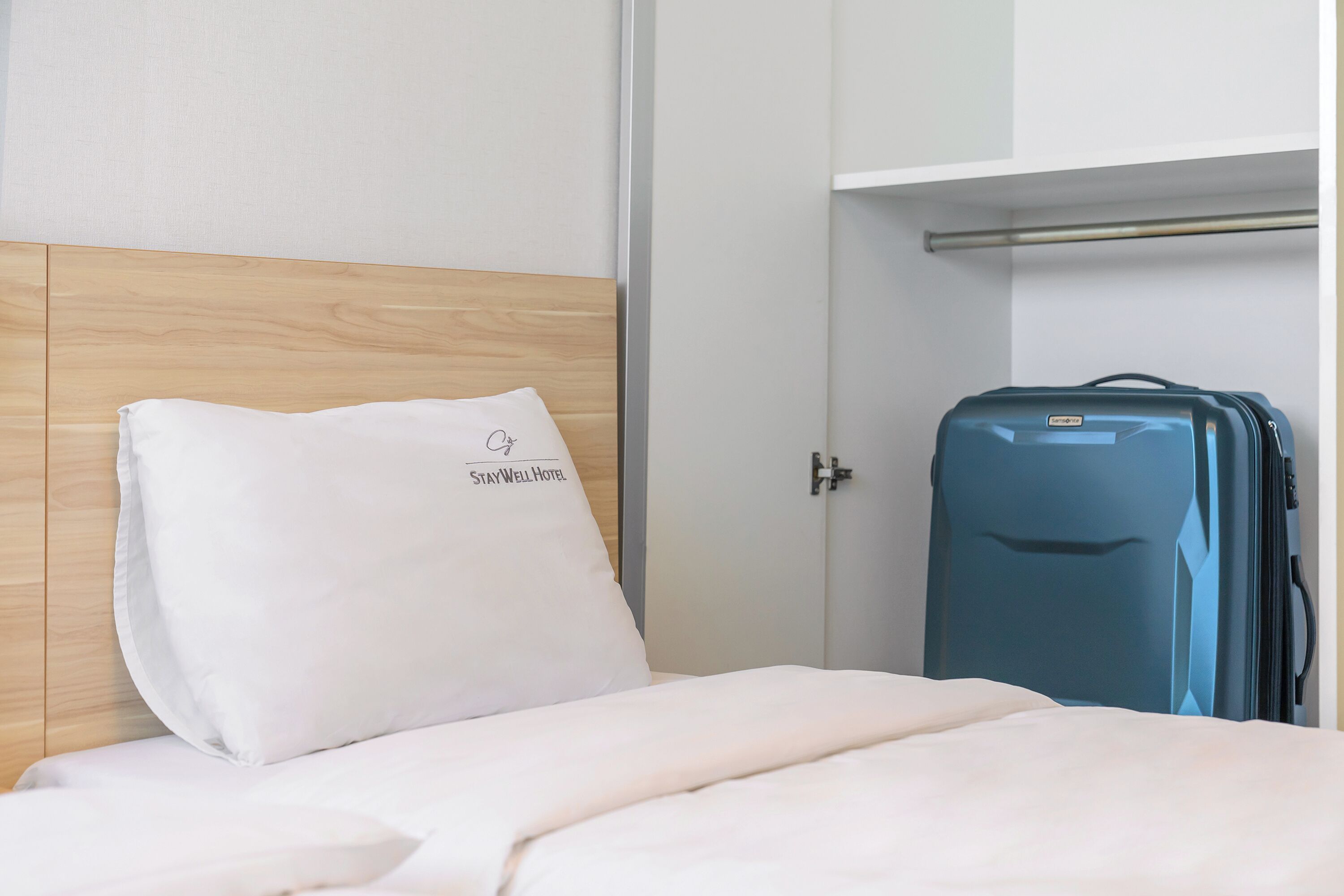 A hotel room with a bed, a white pillow with a logo, a blue suitcase, and a wardrobe.