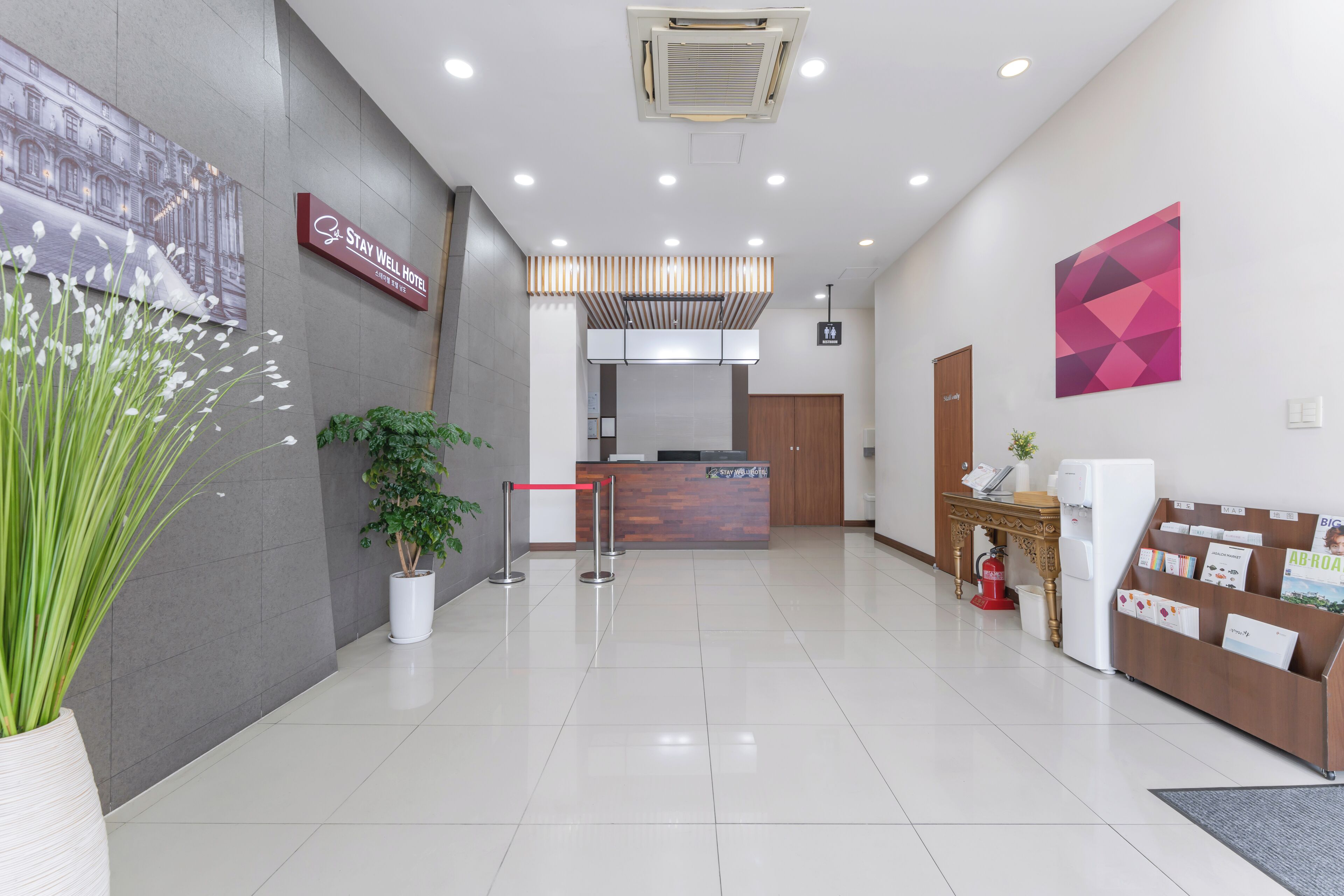 Zona de recepción de hotel con mostrador, una barrera roja, extintor, planta y cartel en la pared.
