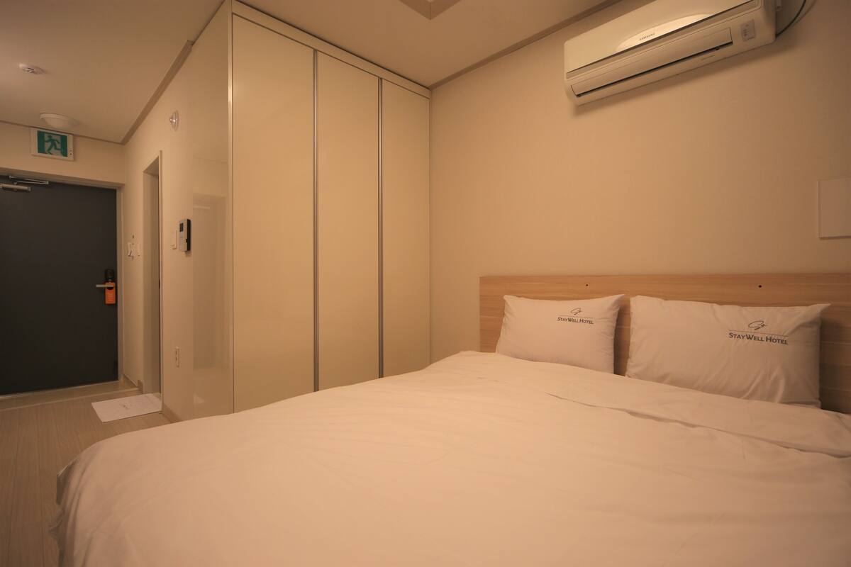 Habitación de hotel con cama, dos almohadas con el logotipo del hotel, un armario y un equipo de aire acondicionado.