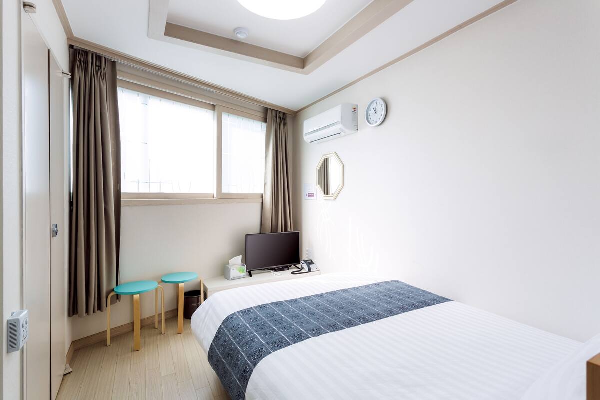Habitación de hotel con cama, escritorio con computadora, ventana con cortinas, aire acondicionado y un reloj de pared.