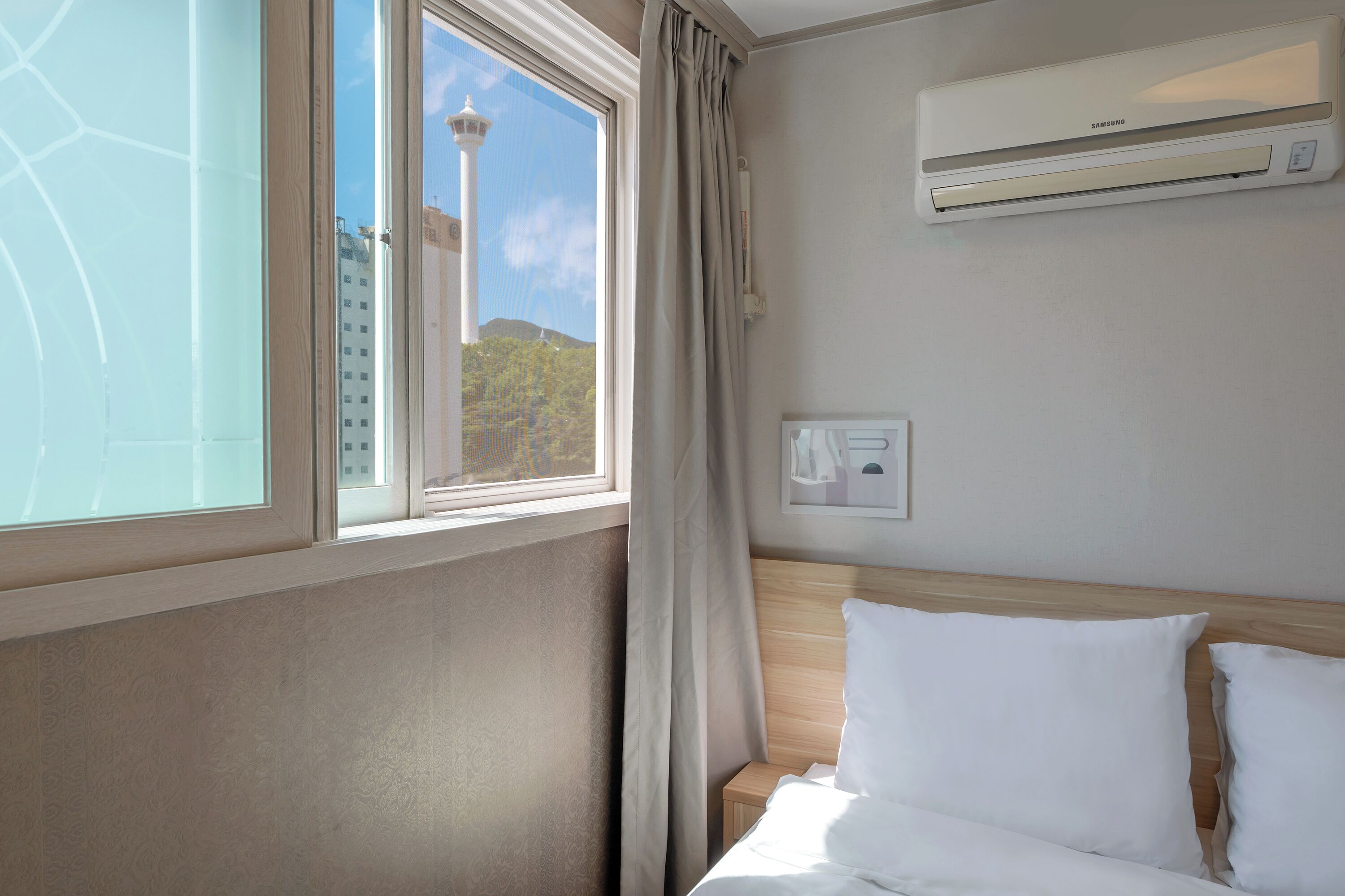Quarto de hotel com cama, ar condicionado, janela com cortinas e vista para uma torre.