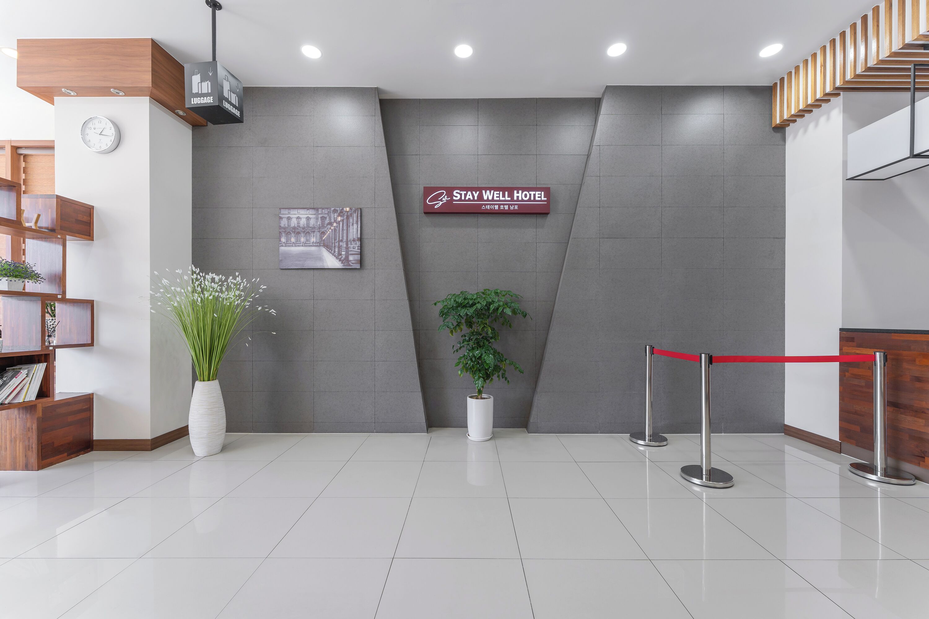 A reception area with a clock, a sign for Stay Well Hotel, a potted plant, and a red rope barrier.