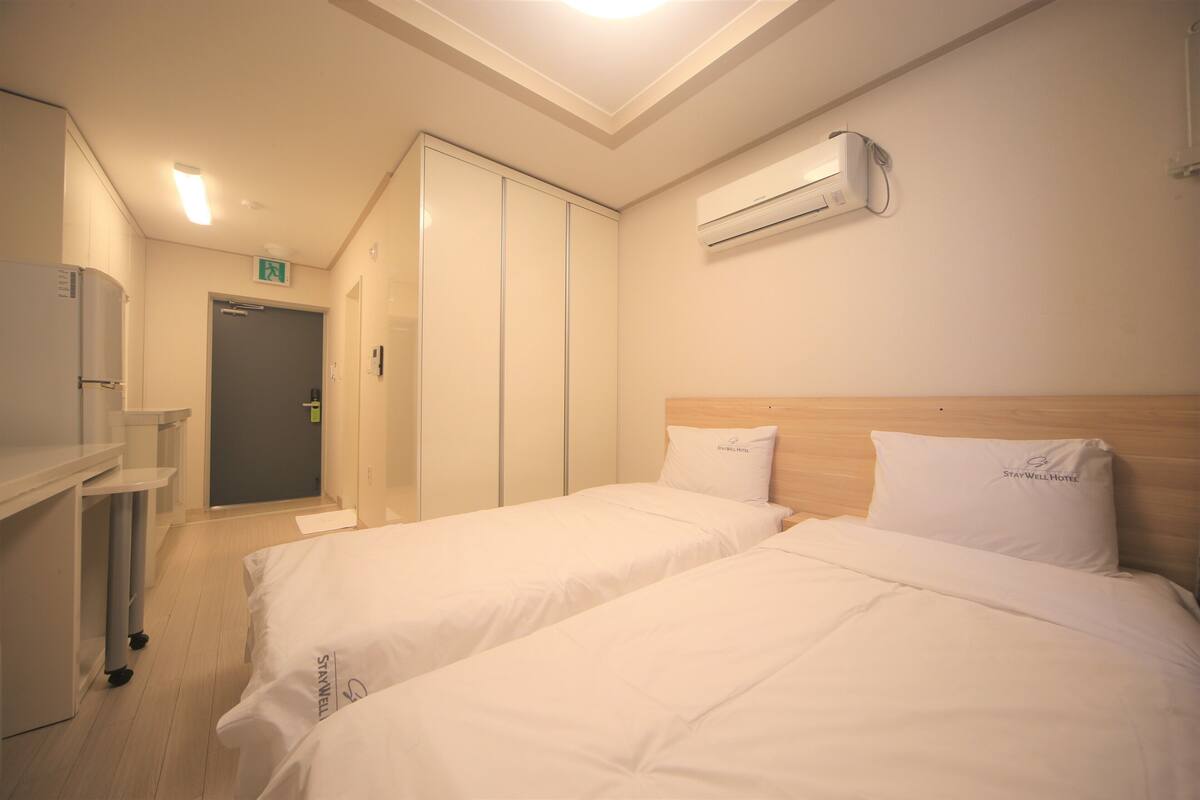 Habitación de hotel con dos camas, un armario, un escritorio y un aire acondicionado.